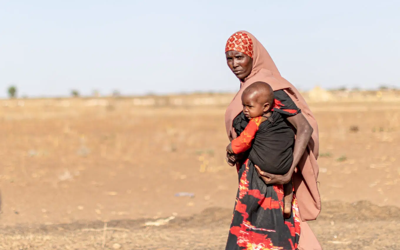 Woman hold child standing in front of brown pasture.