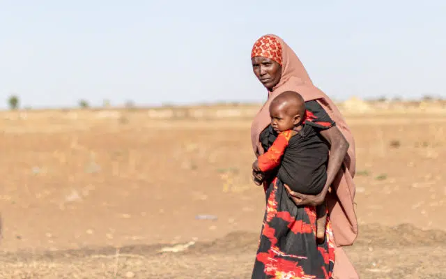 Woman hold child standing in front of brown pasture.