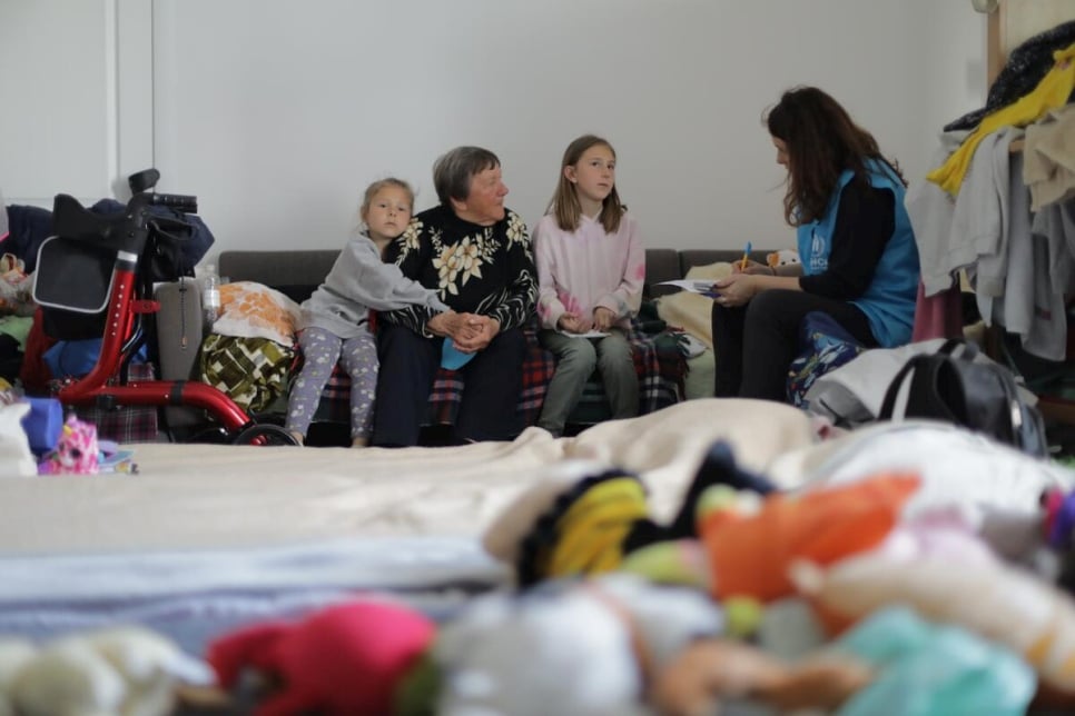 An older woman with short grey hair sits between two younger girls on a couch, the one on her left is the youngest and is clinging onto her arm, all three are speaking to a female UNHCR worker on the far right 