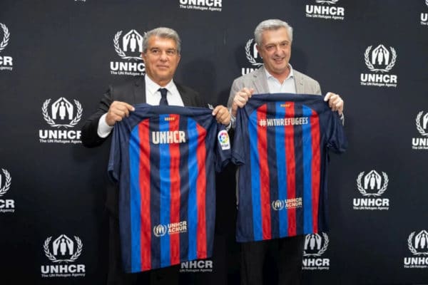 two men holding up soccer jerseys that have black, blue, and red vertical stripes and say UNHCR in white writing centred at the neck line on the back of the jerseys