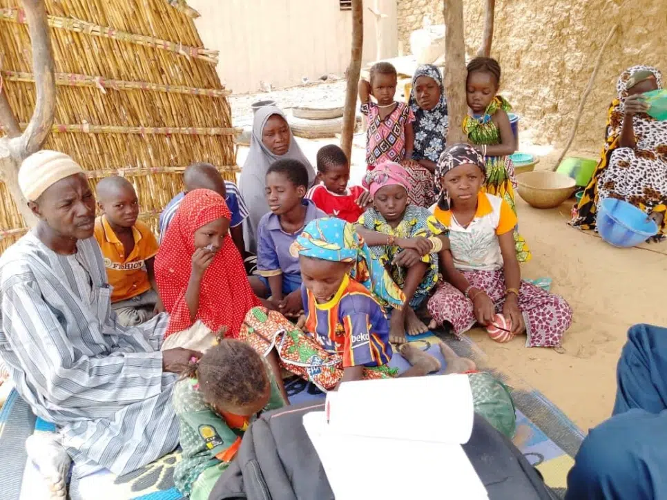 group of kids, varying ages, sitting on sand in front of straw walls