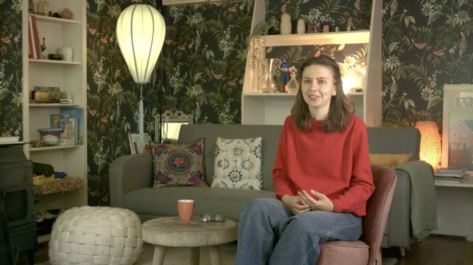 Woman wearing red long sleeve and grey pants sitting on chair in a living room with olive coloured, floral, wallpaper 