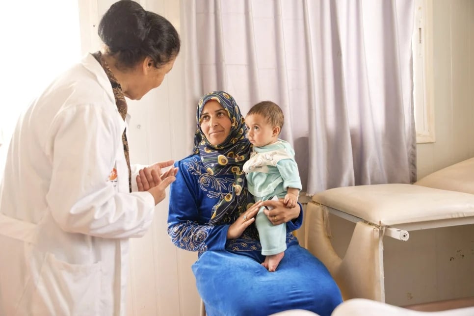 An Arab female doctor is speaking to an Arab woman wearing a blue dress and a blue patterned head scarf, while the woman is holding a baby. They are in a doctors office. 