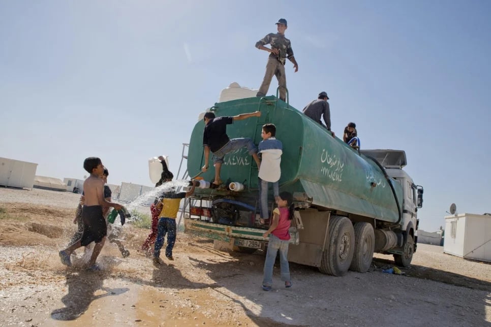 Children play with water coming out of a water truck while three men stand smiling at the top of the truck looking down on them