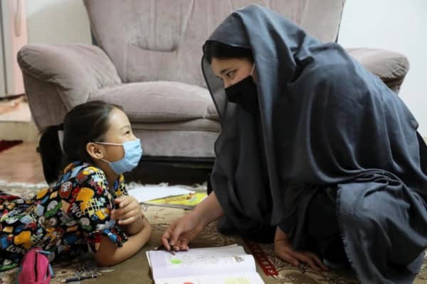 A woman wearing a dark head scarf and a black mask is smiling and kneeling in front of a young girl who is wearing a medical mask laying on her stomach looking up at her