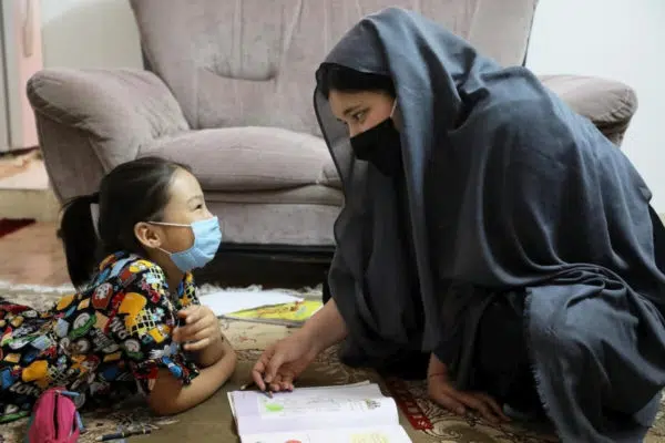 A woman wearing a dark head scarf and a black mask is smiling and kneeling in front of a