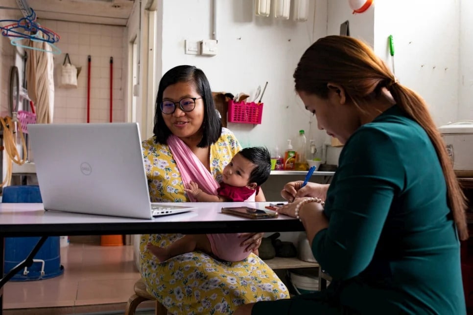 Two asian women sitting at a table, across from each other, the one on the left of the frame is looking at a laptop while holding a baby, while the woman on the right of the screen is writing on paper