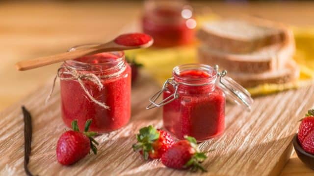 glass jars filled with red jam on a table in front of couple pieces of bread