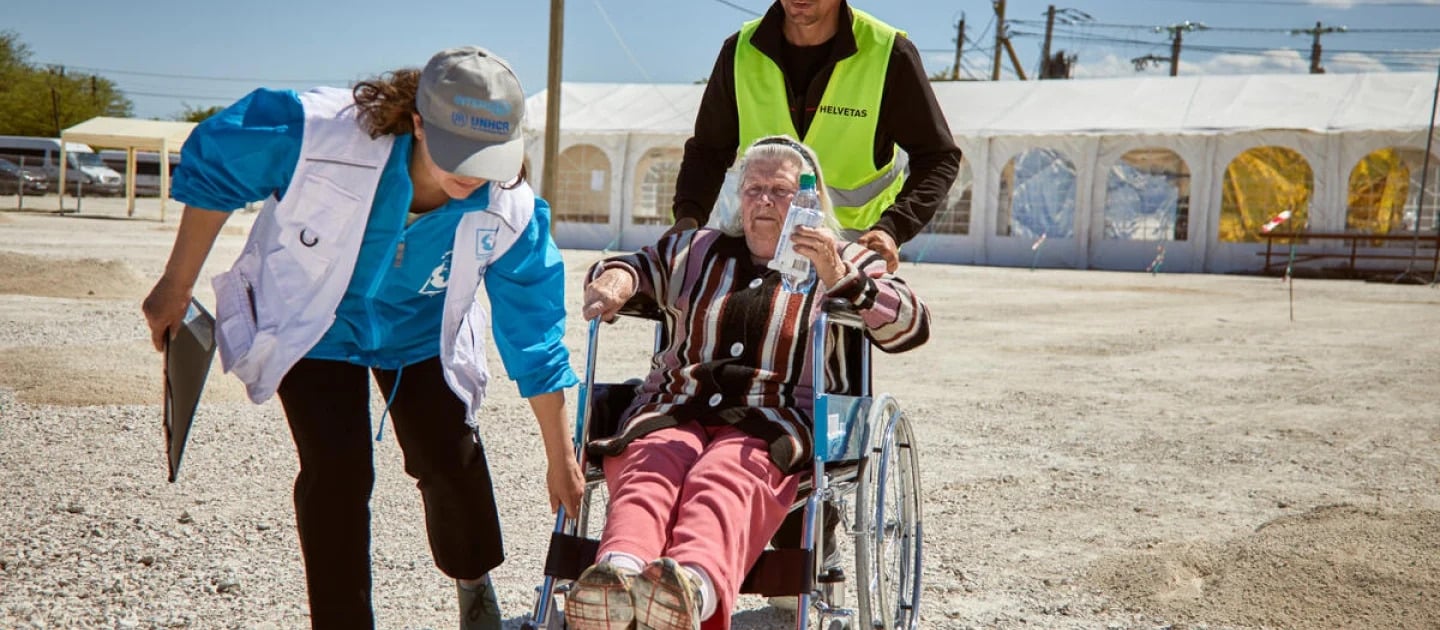 UNHCR partner wearing a blue long-sleeve shirt under a white vest helps slightly lift the wheelchair of an older woman who is sitting in it wearing red pants and a vertically stripped shirt, while another person wearing a black long-sleeve under a reflective vest pushes the wheel chair from behind