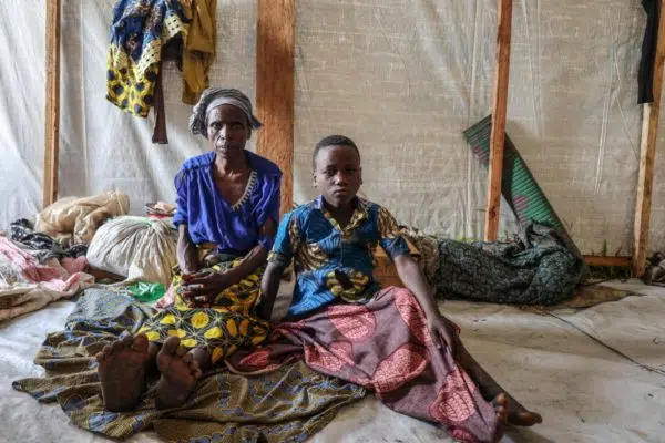 An older black woman wearing a head wrap and a younger boy sitting next to each other on the ground wearing bright coloured, patterned, clothing with a grim look on their faces