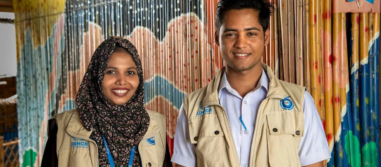 A woman, wearing a dark patterned head scarf, is standing next to a man, both are wearing UNHCR khaki vests and standing in front of a wall of coloured bamboo 