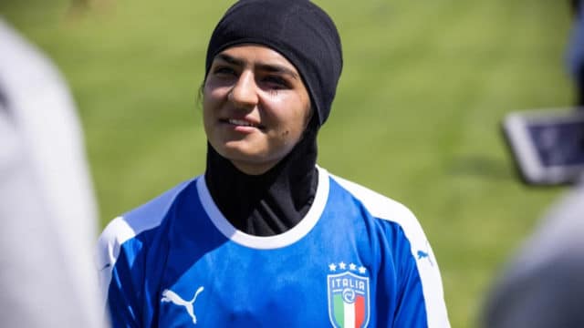woman wearing a head scarf and an Italian soccer jersey looks off into the distance behind the camera