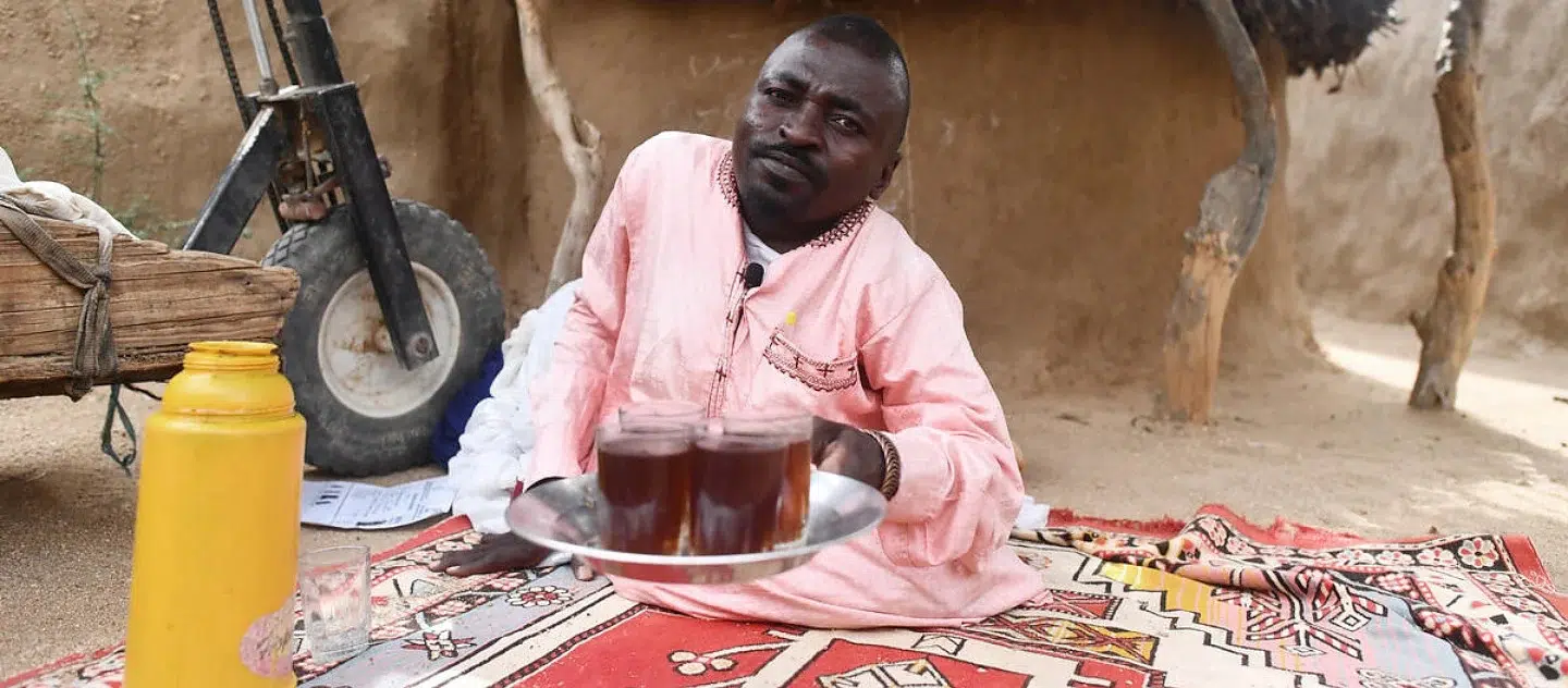 Man without legs sits on a blanket in sand holding a tray with four glasses on it filled with liquid