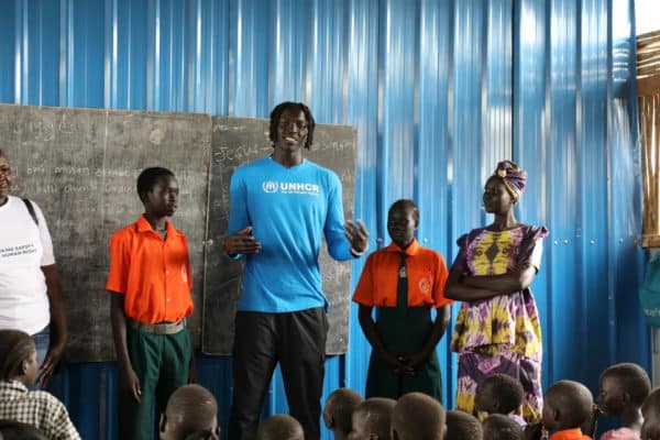 Tall black man wearing a UNHCR blue long sleeve is standing in front of a chalkboard speaking to a classroom of South Sudanese children
