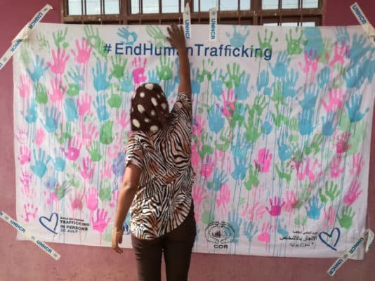 A black woman wearing a head scarf is standing in front of a wall with a hanging white sheet which is covered in pink, blue, and green handprints. The top of the sheet reads "#EndHumanTrafficking" and the woman is reaching for the top of the sheet near a window.