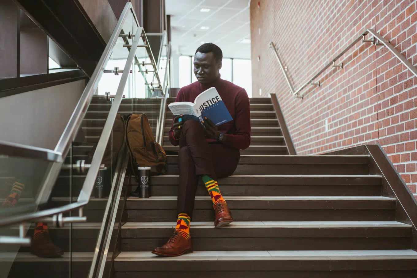 Man sitting on steps of a stairwell reading a white and blue book that says "Justice" on the cover.