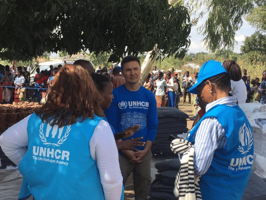 Group of four people standing in circle. They are wearing blue vests with UNHCR logos standing in front of a line of people in the back.