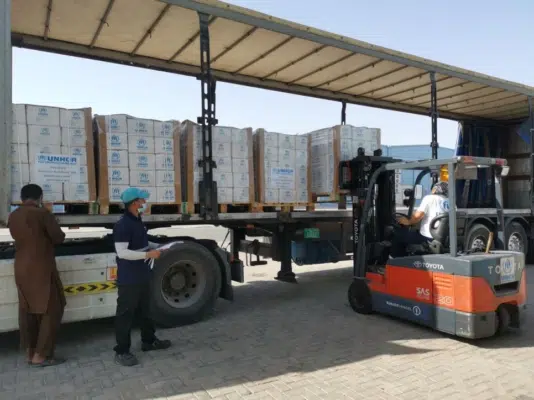 Freight truck with UNHCR boxes of emergency supplies. To the left there is someone operating a forklift adding a palette of boxes onto the truck.