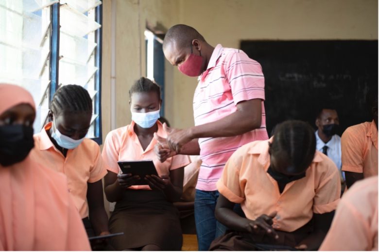 School youth in pink uniform with masks on sitting in classroom. Teacher stands over student with tablet in her hand.