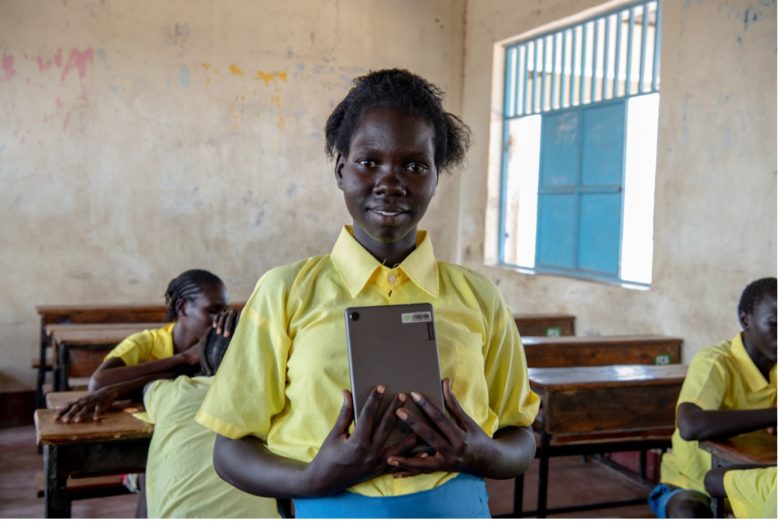 Youth in yellow button up school uniform standing in classroom holding a tablet by her chest.