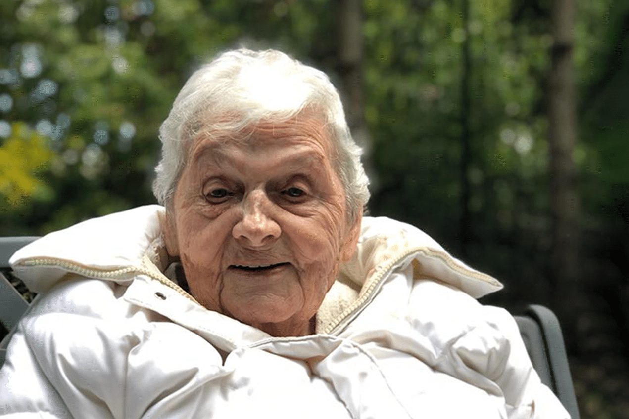 Eldery woman with short hair in white puffer jacket sitting outdoors in front of trees with the sun shining behind her.