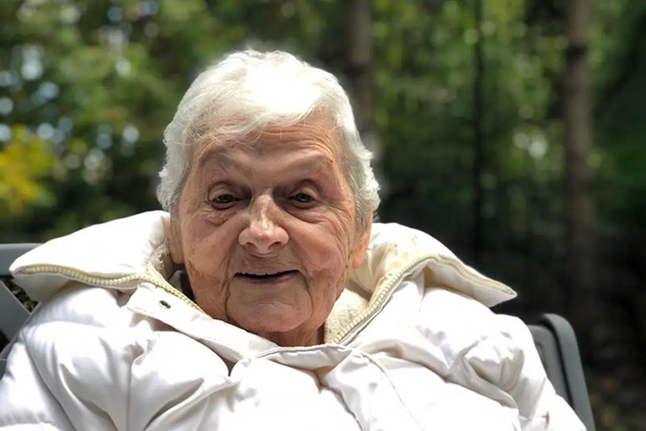 Eldery woman with short hair in white puffer jacket sitting outdoors in front of trees with the sun shining behind her.