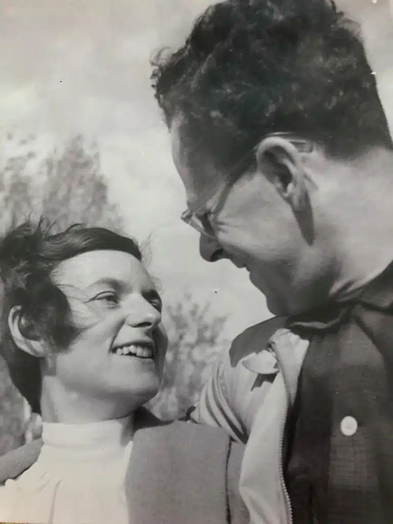 Man on right with arm over the shoulder of a woman with short pixie cut facing each other smiling.