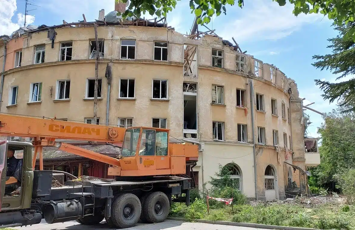 Round beige building with roof collapsed. The windows all have windowpanes shattered or missing. There is a construction truck in front.