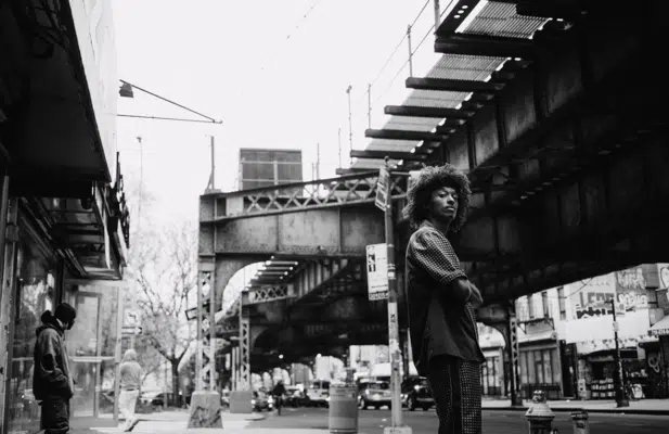 Black and white photo of man standing outside near by a bridge.