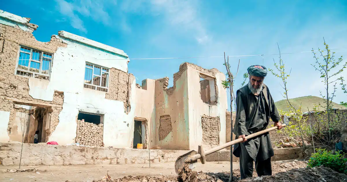 Man stand in front of partially destroyed building with windows bricked in. He is holding a shovel.