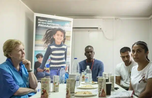 Woman in blue UNHCR vest sits on the left side of the table across from three people.