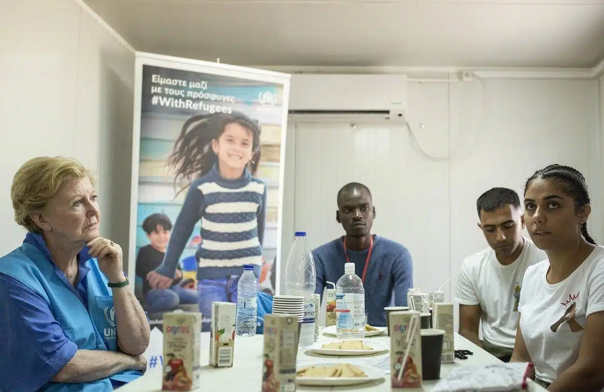 Woman in blue UNHCR vest sits on the left side of the table across from three people. There is a banner with a UNHCR logo and child at the back.