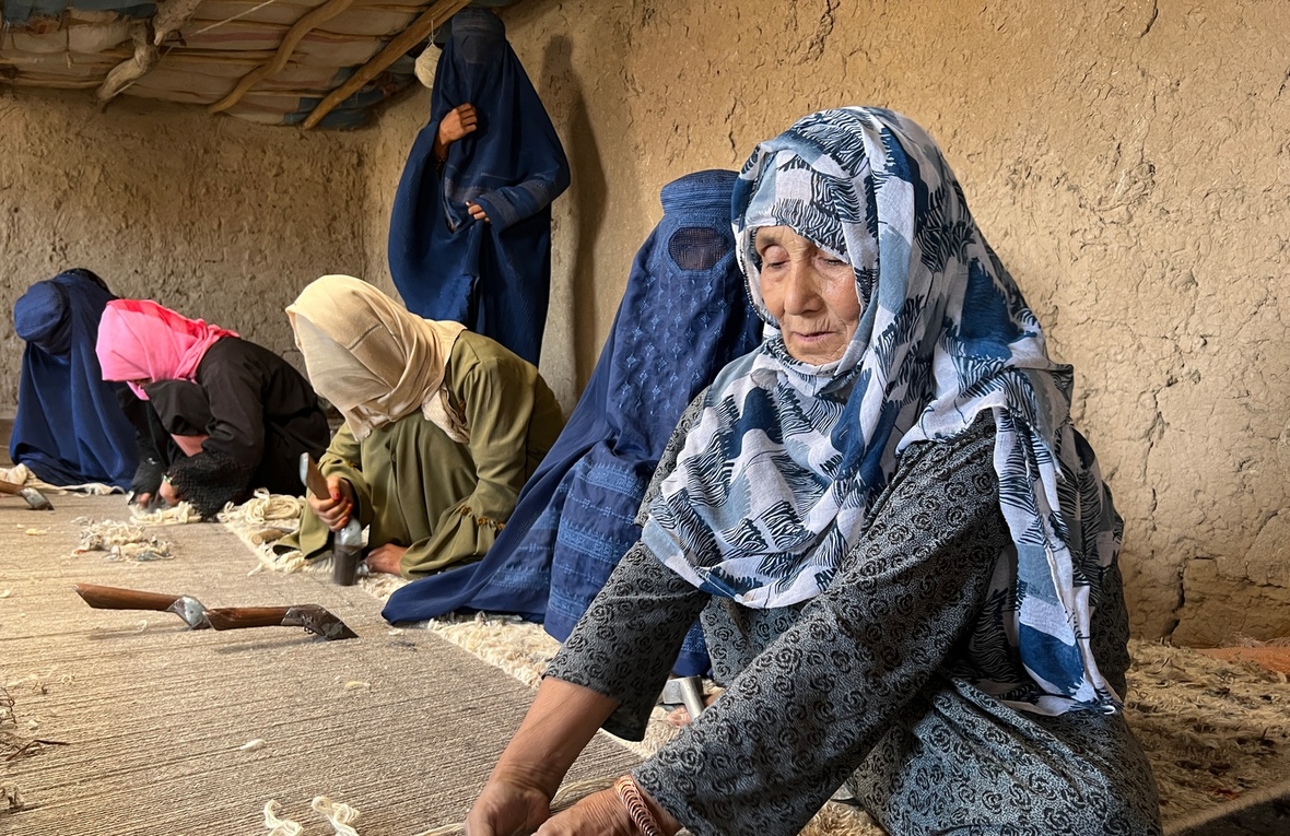 Elderly woman with blue and white head scarf sits on the floor with wool in her hand.