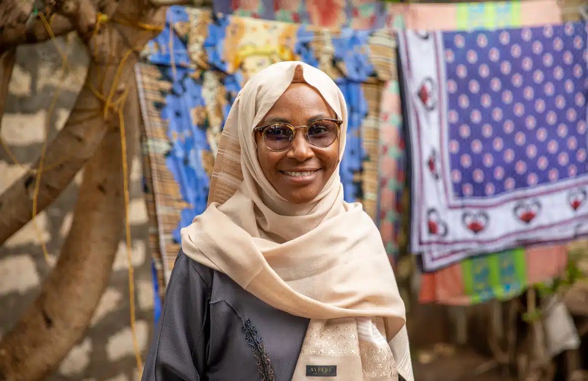 Woman with beige coloured head scarf with sunglasses on smiles.