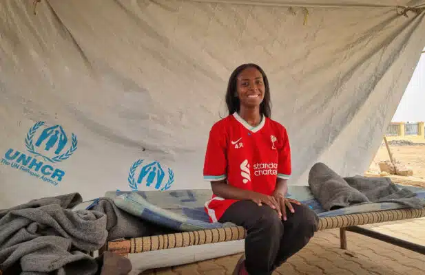 Woman in red shirt and black pants sitting on a bench in front of a tarp in the back with the UNHCR logo.