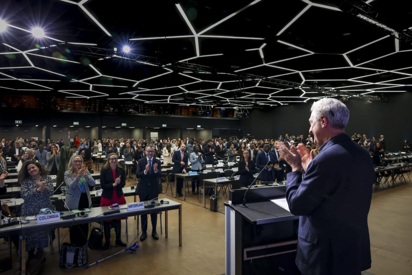 UN High Commissioner for Refugees Filippo Grandi stands in front of a podium, clapping, in a room with a black geographic cieling an audience of world leaders. 
