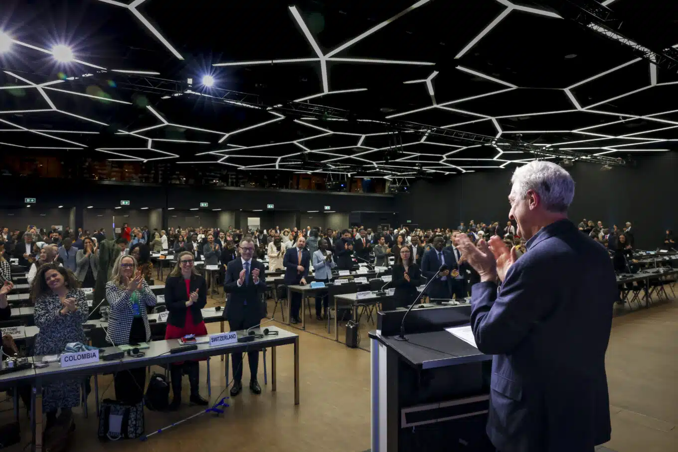 UN High Commissioner for Refugees Filippo Grandi stands in front of a podium, clapping, in a room with a black geographic cieling an audience of world leaders. 