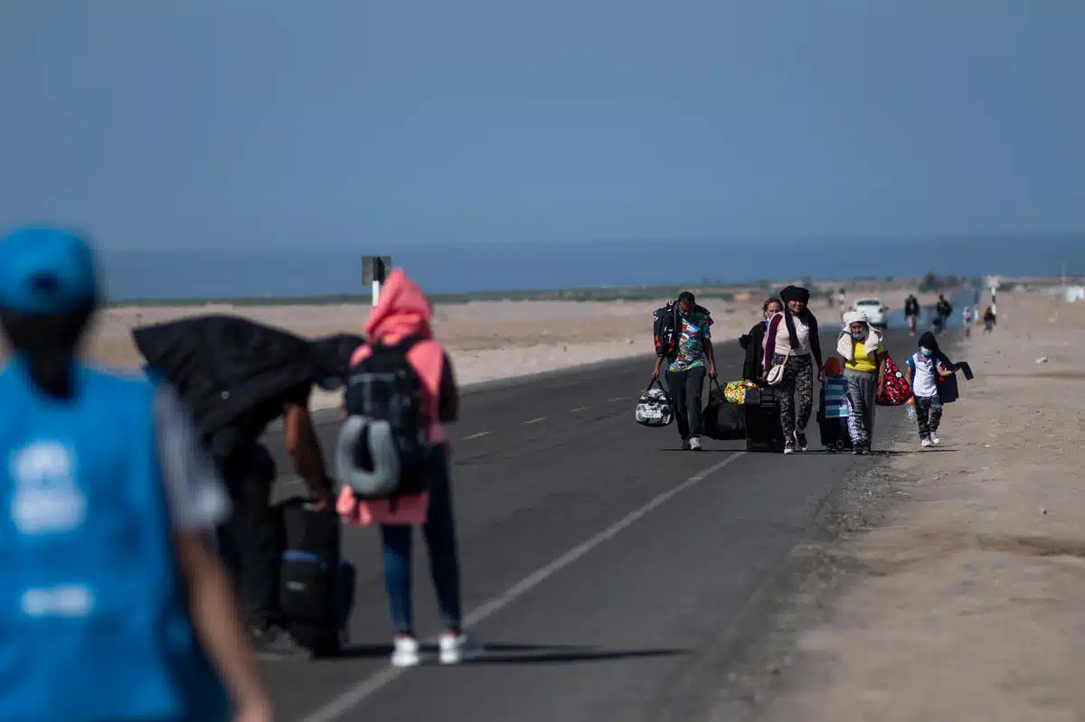 a group of six people walk on a road in the desert-like area carrying bags. 