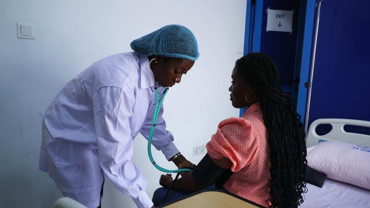 A young woman dressed in a doctor's whote coat and her heir in a blue hairnet listens to a patient's heart with a stethescope. They are in a hispital room with a dark blue door. 