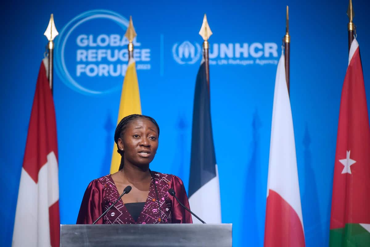 une jeune femme se tient derrière un podium devant cinq drapeaux de pays sur un fond bleu du forum mondial des réfugiés 