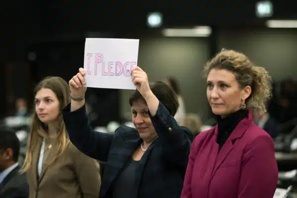 A woman in a blazer holds up a piece if paper with the words "I pledge" written on it in pink bubble letters.