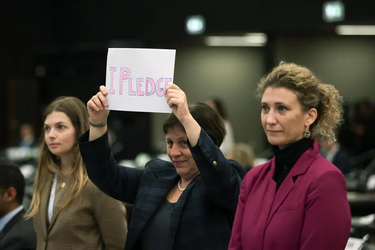 Une femme vêtue d'un blazer brandit un morceau de papier sur lequel sont inscrits les mots "I pledge" (je m'engage) en lettres roses à bulles. 