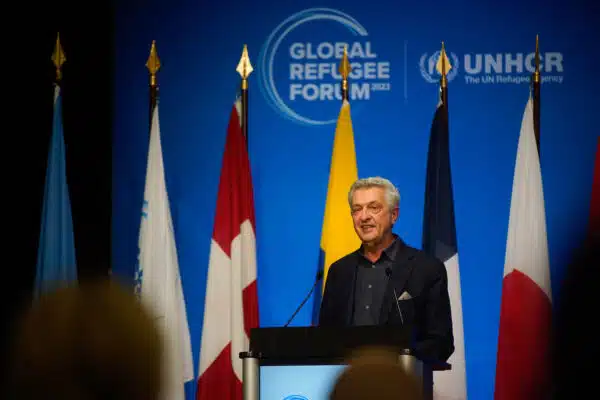 UN High Commissioner for Refugees Filippo Grandi stands behind a podium, in frony of four country flags and a blue global refugee fourm backdrop