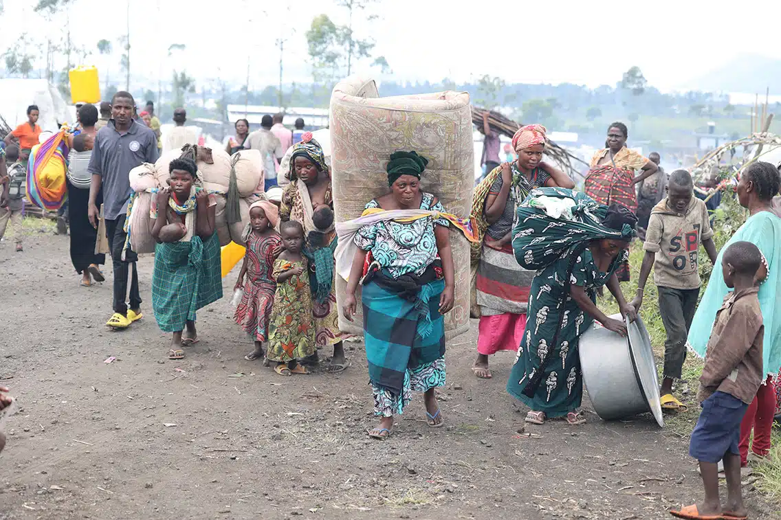 A group of people walk along a dirt road. Manny carry cloth bags and a woman in the middle has a mattress tied to her back. 