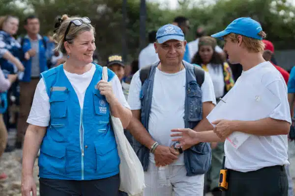 three people stand talking to each other, one wearing a unhcr vest and two wearing unhcr caps