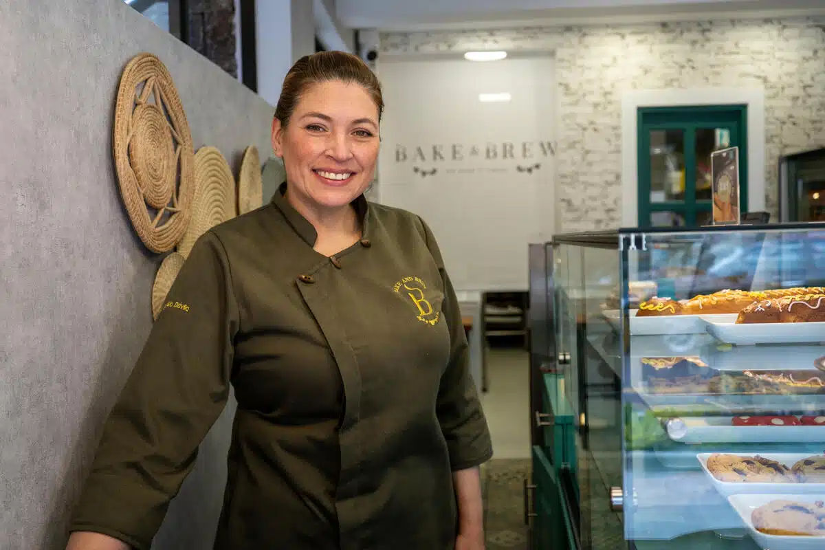 A woman smiles wearing a chef's coat in front of a pastry case and dining area