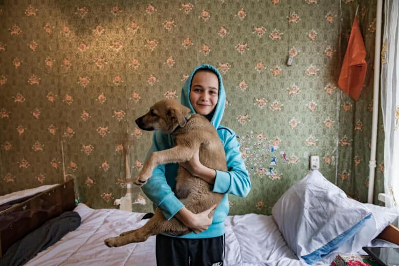 A boy in a light blue hoodie holds a young puppy in a bedroom.