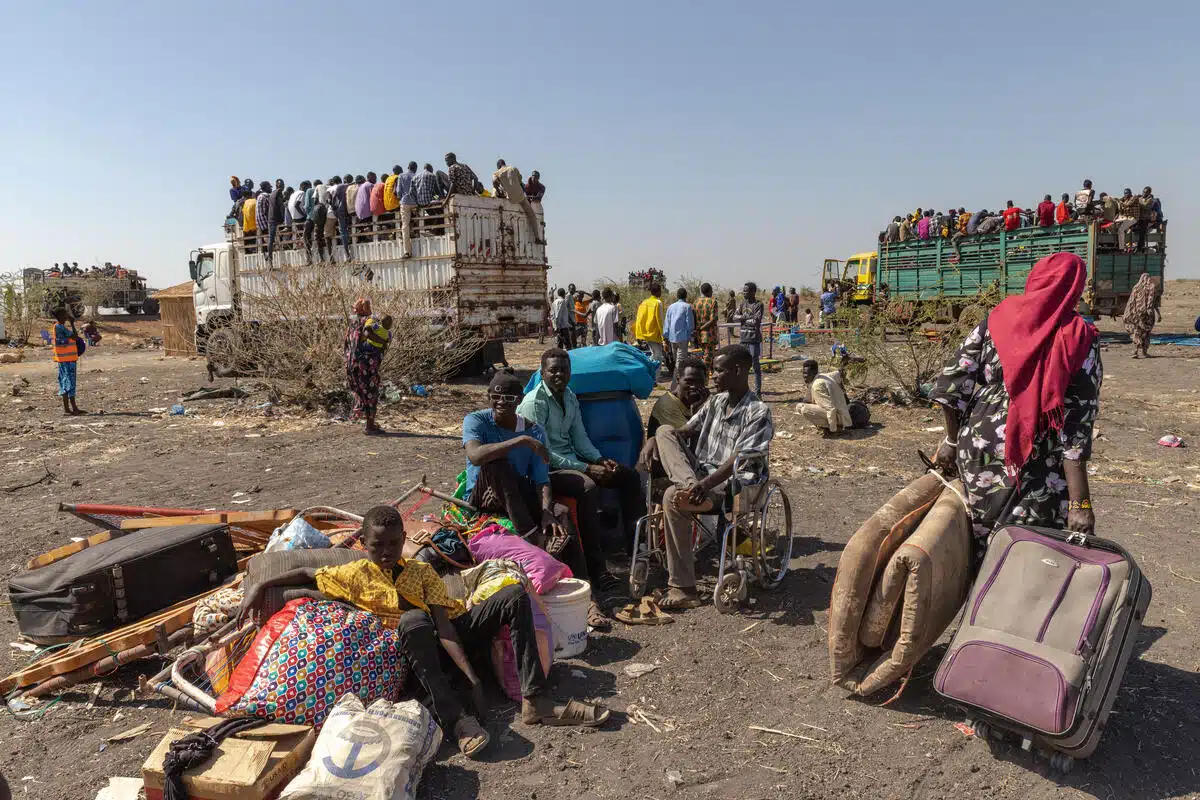 Cinq personnes, dont l'une est en fauteuil roulant, sont assises au milieu de matelas et de sacs devant une foule de personnes assises dans des camions. 