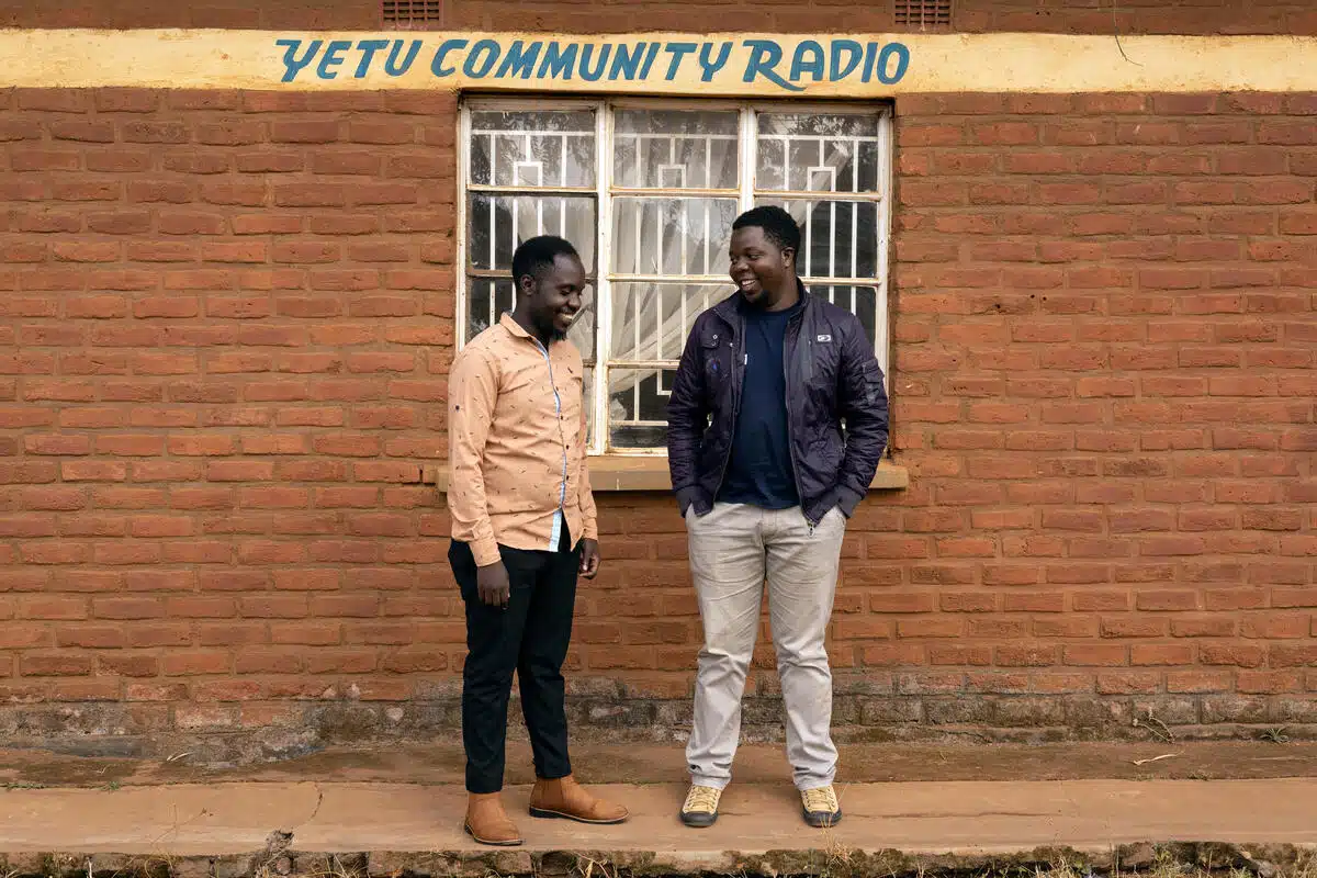 Two men stand in front of a brick wall with the words "yetu community radio" written on the wall above them 