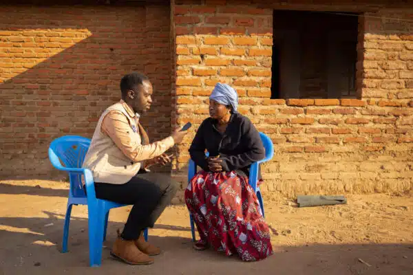 A man holding a voice recorder interviews a woman as they bith sit on blue plastic chairs.